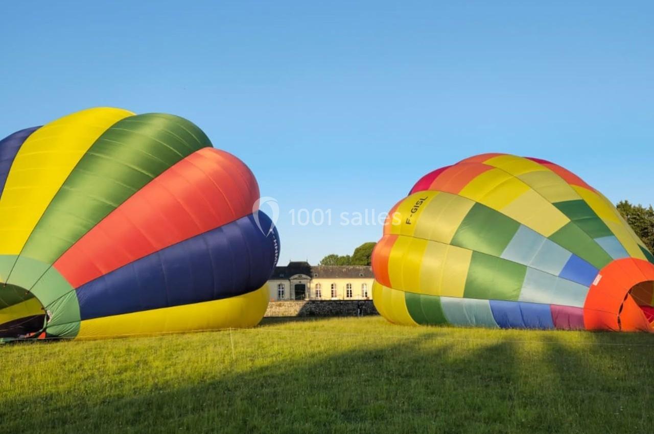 Deux montgolfières colorées en cours de gonflage sur une prairie, avec un bâtiment en arrière-plan sous un ciel bleu.