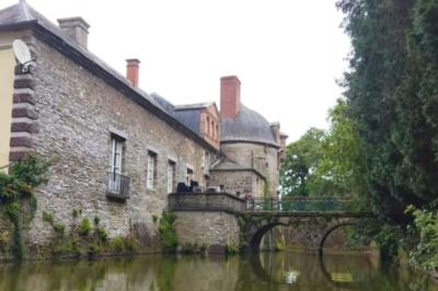 Façade en pierre d'un château bordé par un cours d'eau, avec un pont en arc et des arbres environnants.