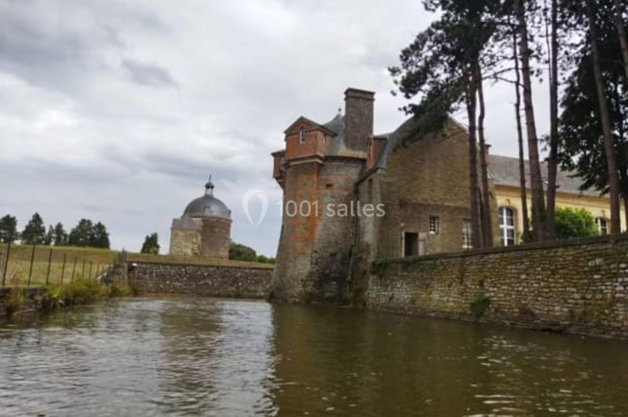 Vue d'un bâtiment historique en pierre près d'un cours d'eau, entouré d'arbres et d'un ciel nuageux.