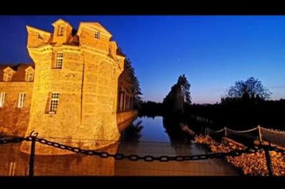 Façade en pierre d'un château bordé par un cours d'eau, avec un pont en arc et des arbres environnants.