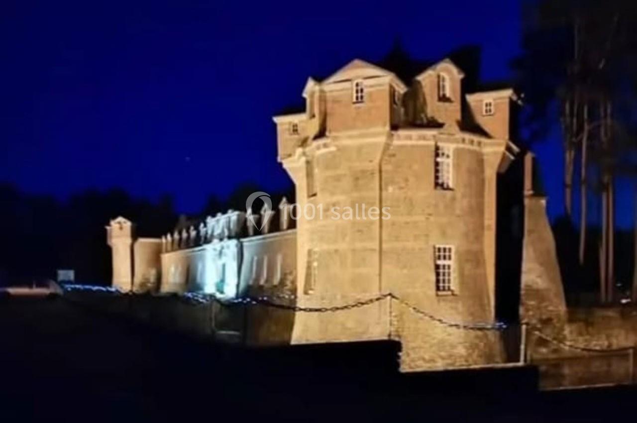 Château en briques éclairé la nuit, entouré de chaînes et d'arbres, avec un ciel bleu foncé en arrière-plan.