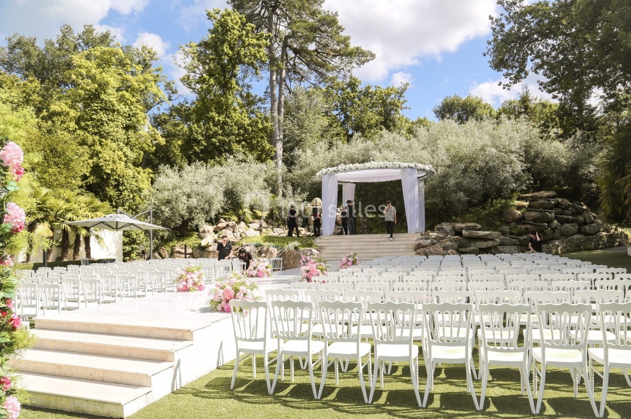Chaises blanches alignées en extérieur face à une arche décorée pour une cérémonie, entourée de verdure.