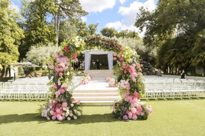 Un photographe capture une cérémonie sous une pergola décorée de fleurs blanches, entourée de verdure et de rochers.