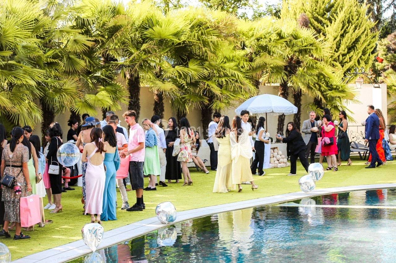 Personnes rassemblées autour d'une piscine dans un jardin, discutant et portant des tenues élégantes sous un ciel ensoleillé.