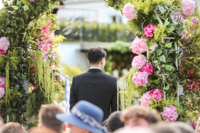 Un photographe capture une cérémonie sous une pergola décorée de fleurs blanches, entourée de verdure et de rochers.