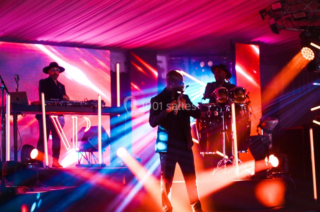 Un musicien joue du saxophone sur scène, accompagné d'un DJ et d'un percussionniste, sous des lumières colorées.