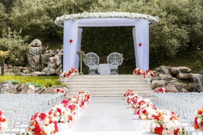 Un photographe capture une cérémonie sous une pergola décorée de fleurs blanches, entourée de verdure et de rochers.