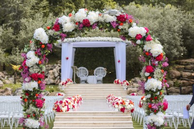 Un photographe capture une cérémonie sous une pergola décorée de fleurs blanches, entourée de verdure et de rochers.