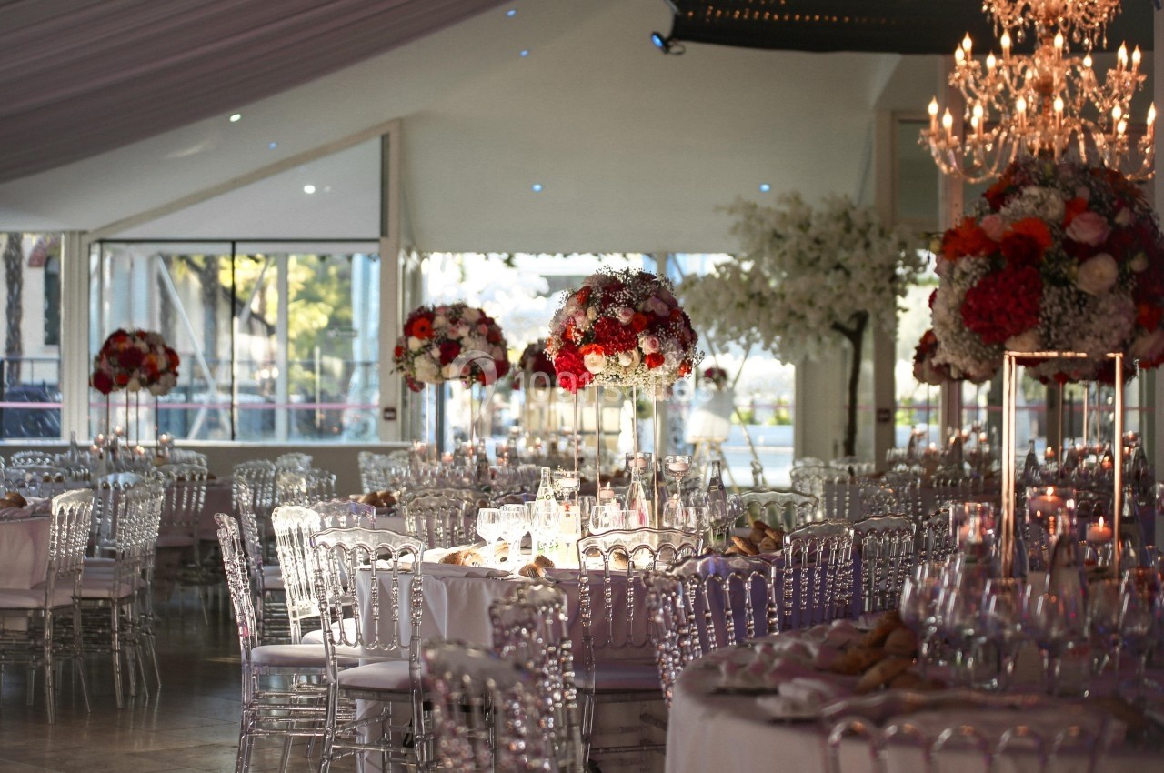 Salle de réception élégante avec tables dressées, chaises transparentes et centres de table fleuris rouges et blancs.