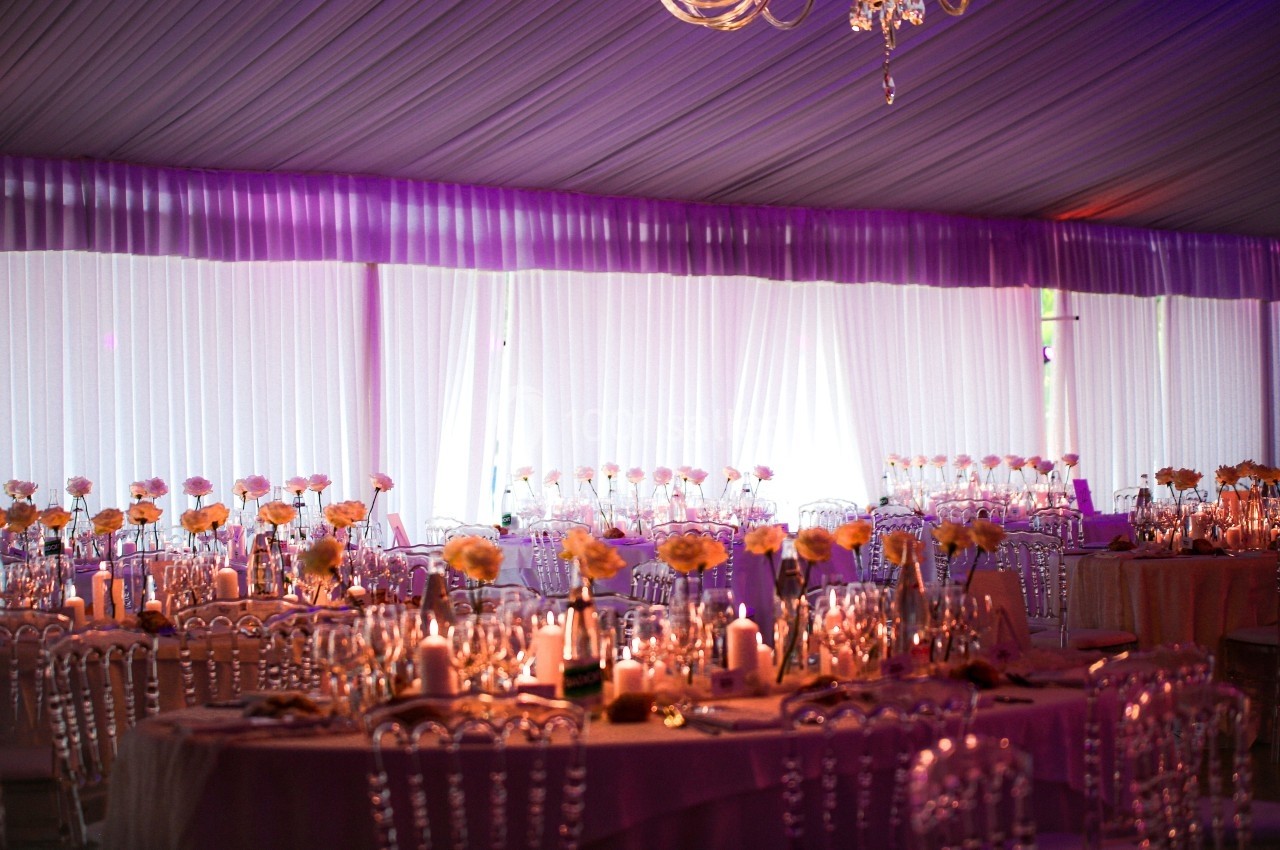 Salle de réception décorée avec des tables rondes, nappes roses, chaises transparentes et centres de table fleuris.