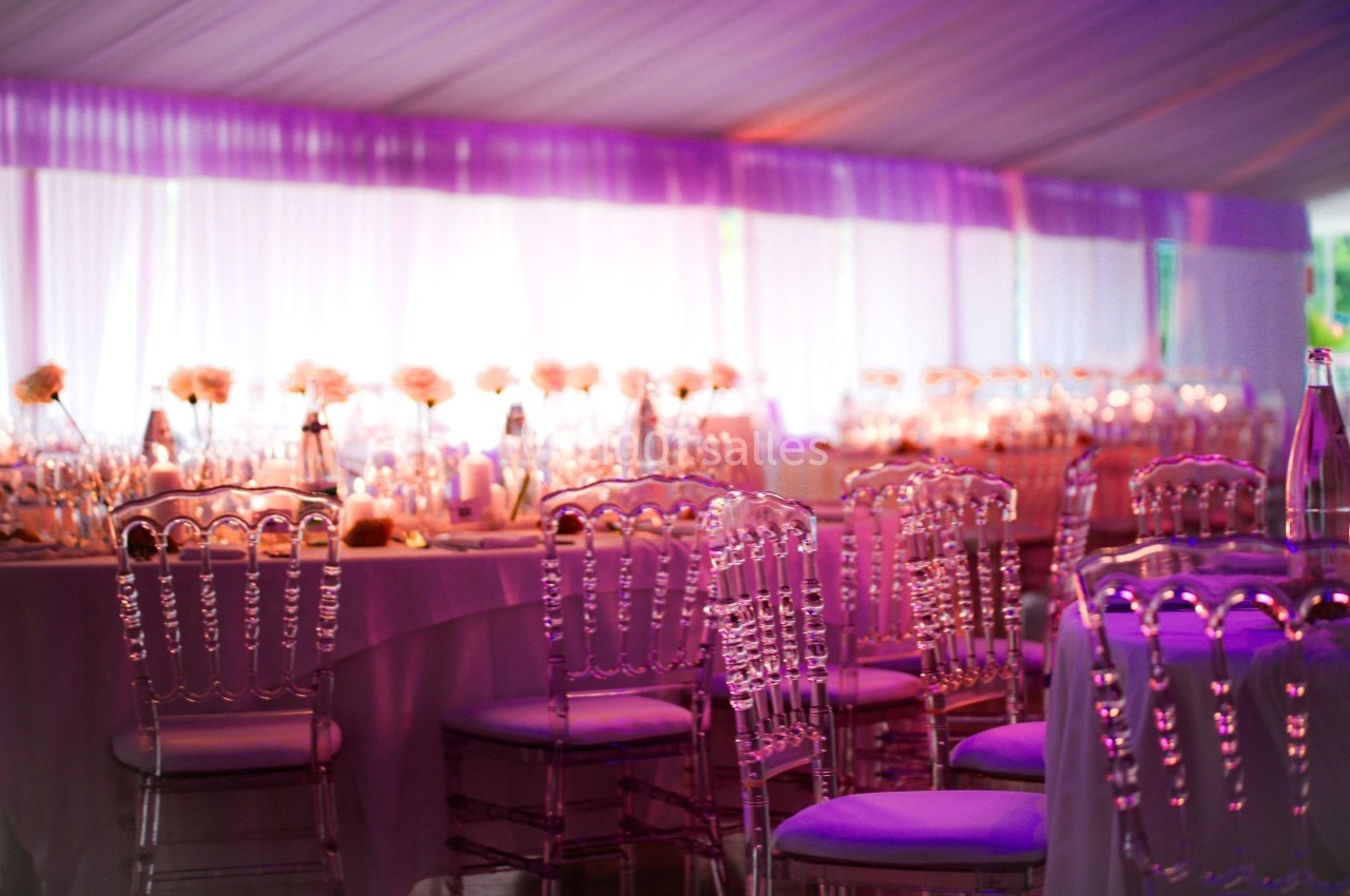Salle de réception décorée avec des chaises transparentes et des tables ornées de fleurs sous un éclairage violet.