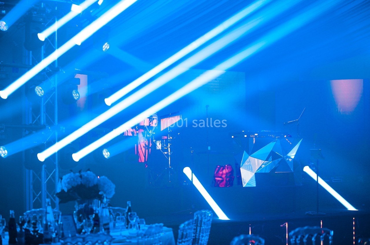 Scène illuminée par des faisceaux lumineux bleus, avec un DJ et un équipement musical en arrière-plan.