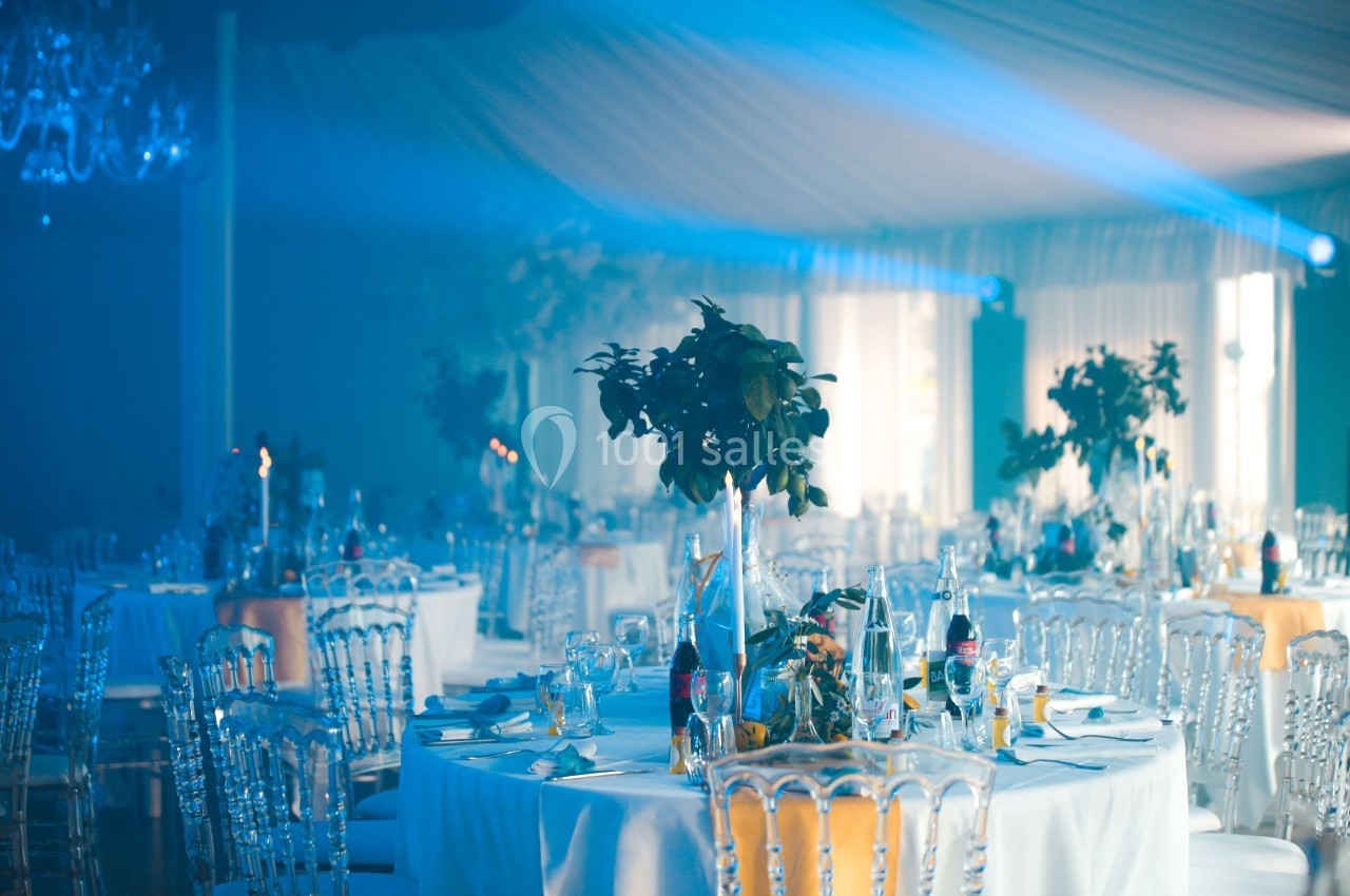 Salle de réception décorée avec des tables rondes, nappes blanches, chaises transparentes et éclairage bleu tamisé.