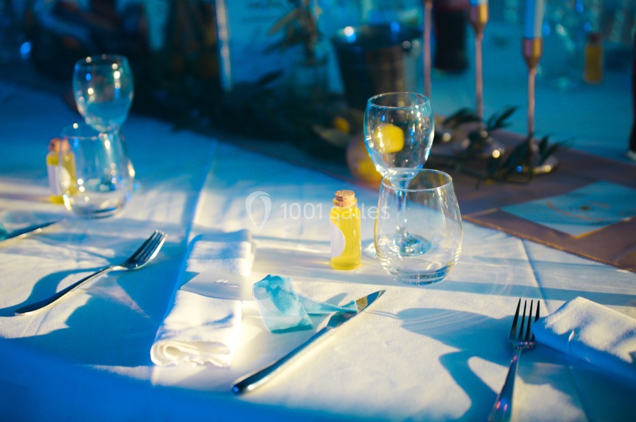 Table dressée avec des couverts, serviettes blanches, verres et petites fioles jaunes sur une nappe blanche éclairée.