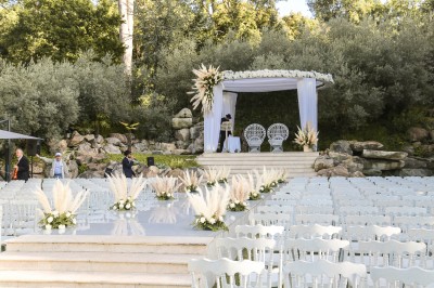 Un photographe capture une cérémonie sous une pergola décorée de fleurs blanches, entourée de verdure et de rochers.