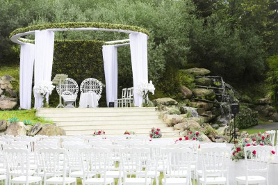 Un photographe capture une cérémonie sous une pergola décorée de fleurs blanches, entourée de verdure et de rochers.