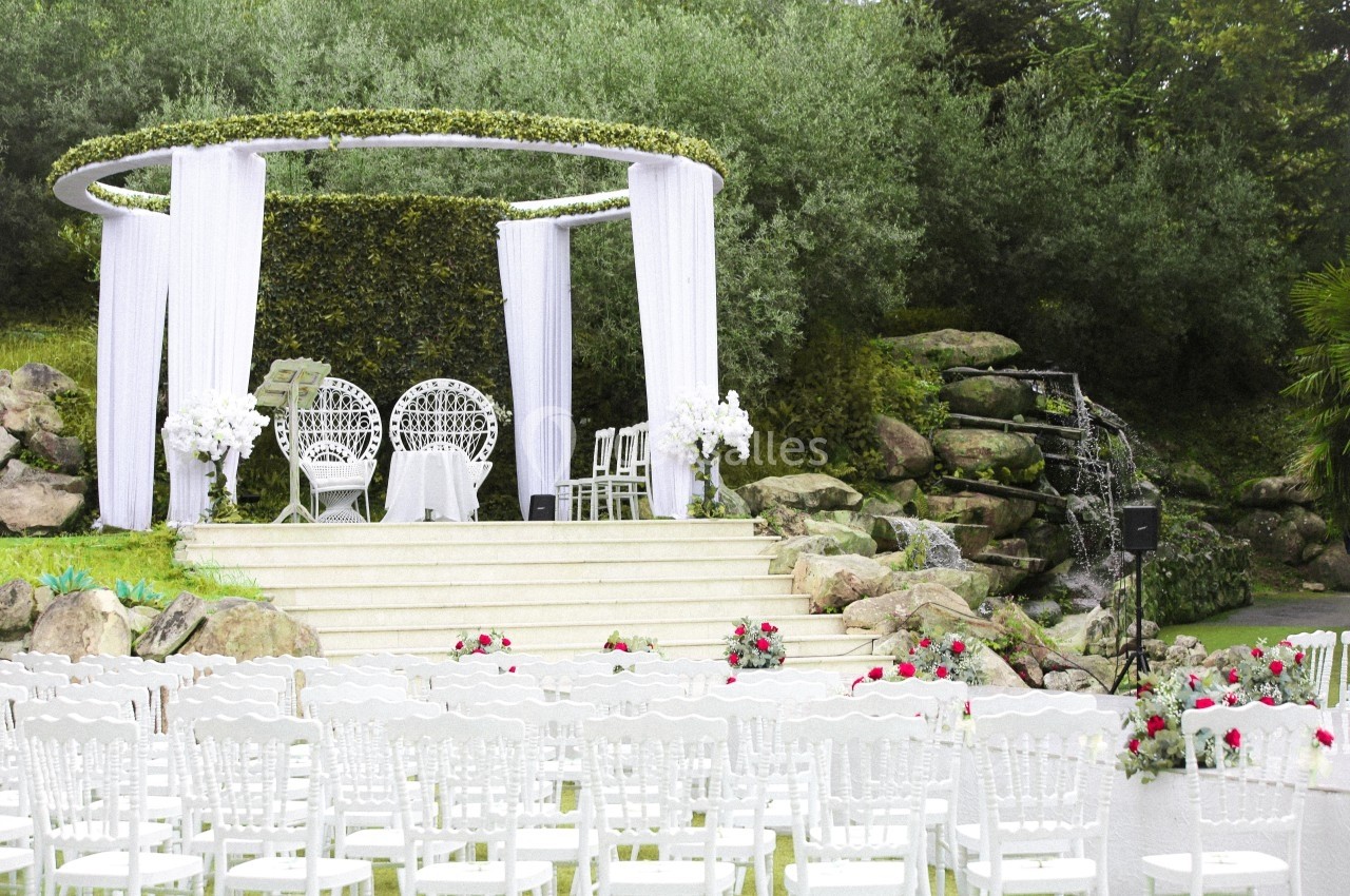 Cérémonie de mariage en extérieur avec arche décorée, chaises blanches alignées et cascade en arrière-plan.