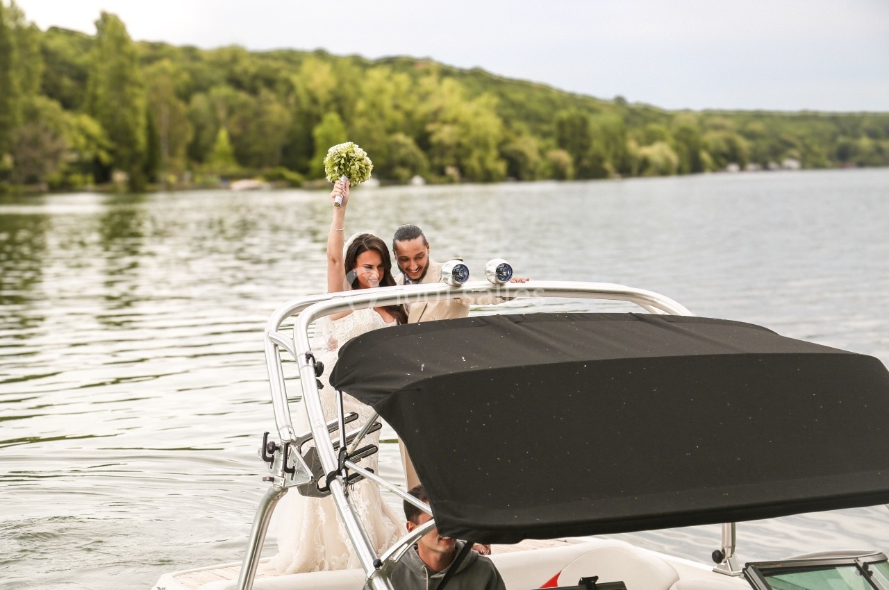 Deux femmes en robe de mariée sur un bateau, l'une levant un bouquet, entourées d'un paysage lacustre verdoyant.