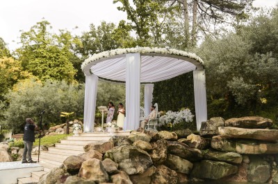 Un photographe capture une cérémonie sous une pergola décorée de fleurs blanches, entourée de verdure et de rochers.
