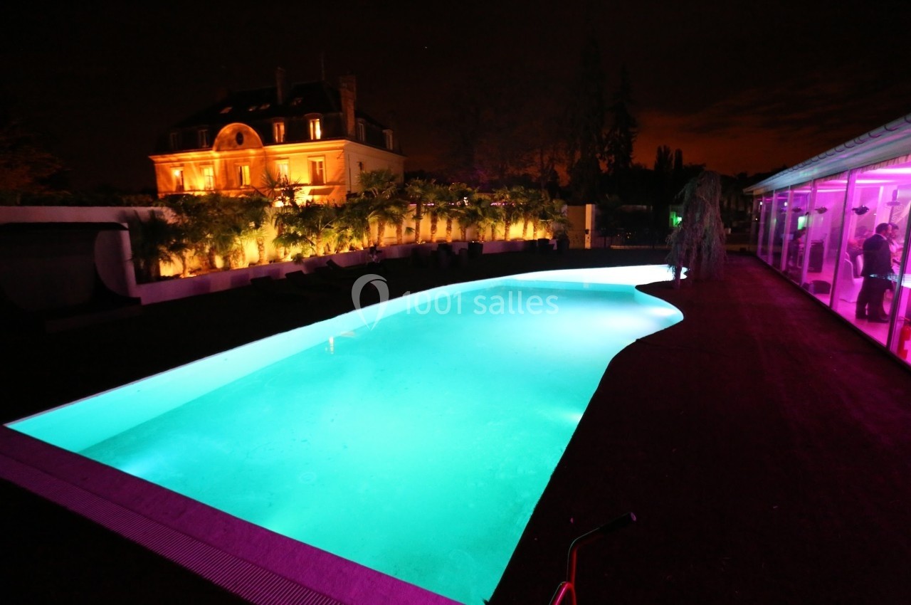Piscine éclairée de nuit devant une grande maison illuminée, entourée de végétation et d'une terrasse.