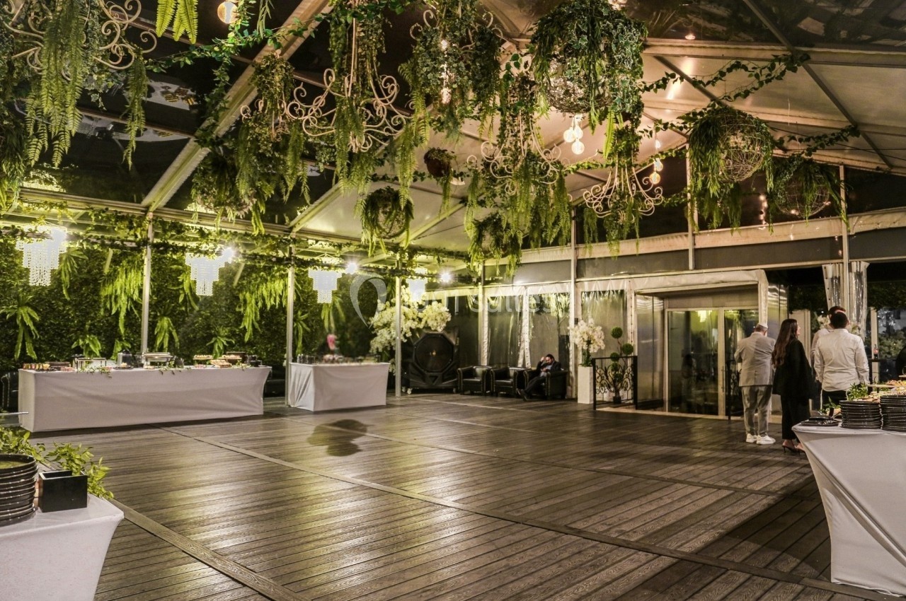 Salle de réception avec parquet, toit transparent, plantes suspendues et tables de buffet décorées.