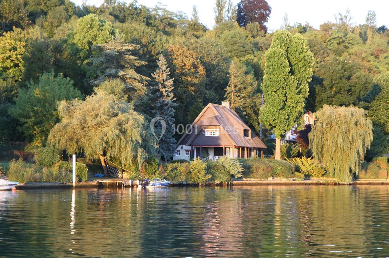 Maison au toit de chaume entourée d'arbres, située au bord d'un lac calme reflétant la végétation environnante.