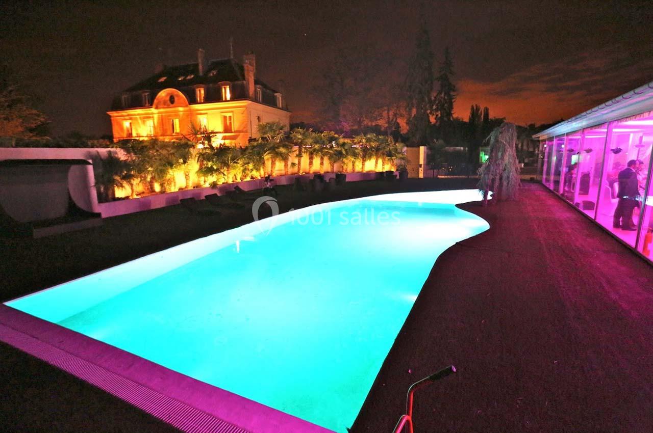 Piscine éclairée de nuit avec une villa illuminée en arrière-plan et des lumières colorées autour.