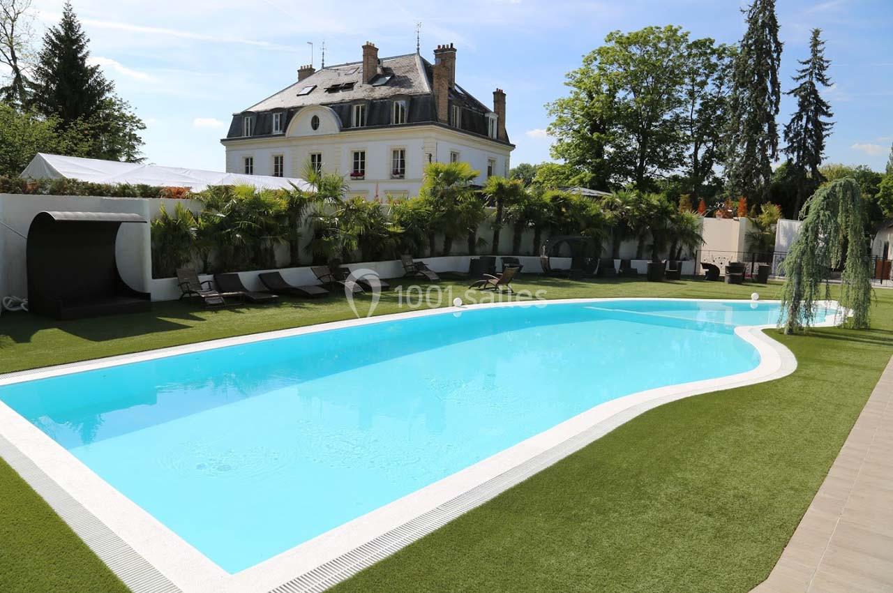 Piscine extérieure entourée de transats, pelouse et végétation, avec un bâtiment de style classique en arrière-plan.