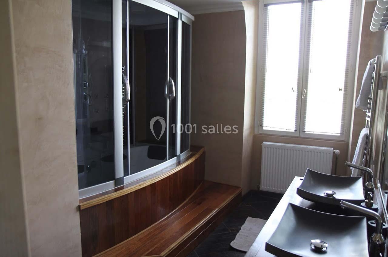 Salle de bain avec une cabine de douche moderne, un banc en bois intégré, deux vasques noires et une fenêtre avec stores.