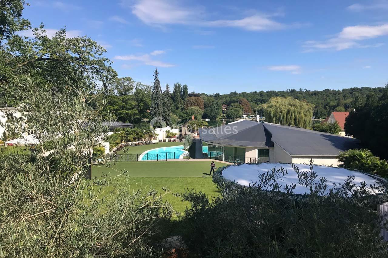 Vue d'un jardin avec piscine, pelouse bien entretenue, bâtiment moderne et arbres en arrière-plan sous un ciel bleu.