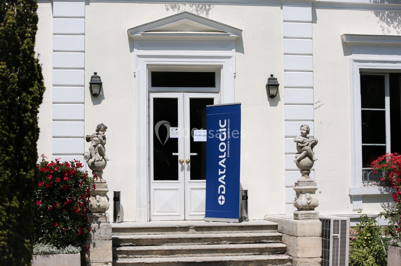 Entrée d'un bâtiment blanc avec une bannière bleue portant le logo ’Datalogic’ près de la porte.