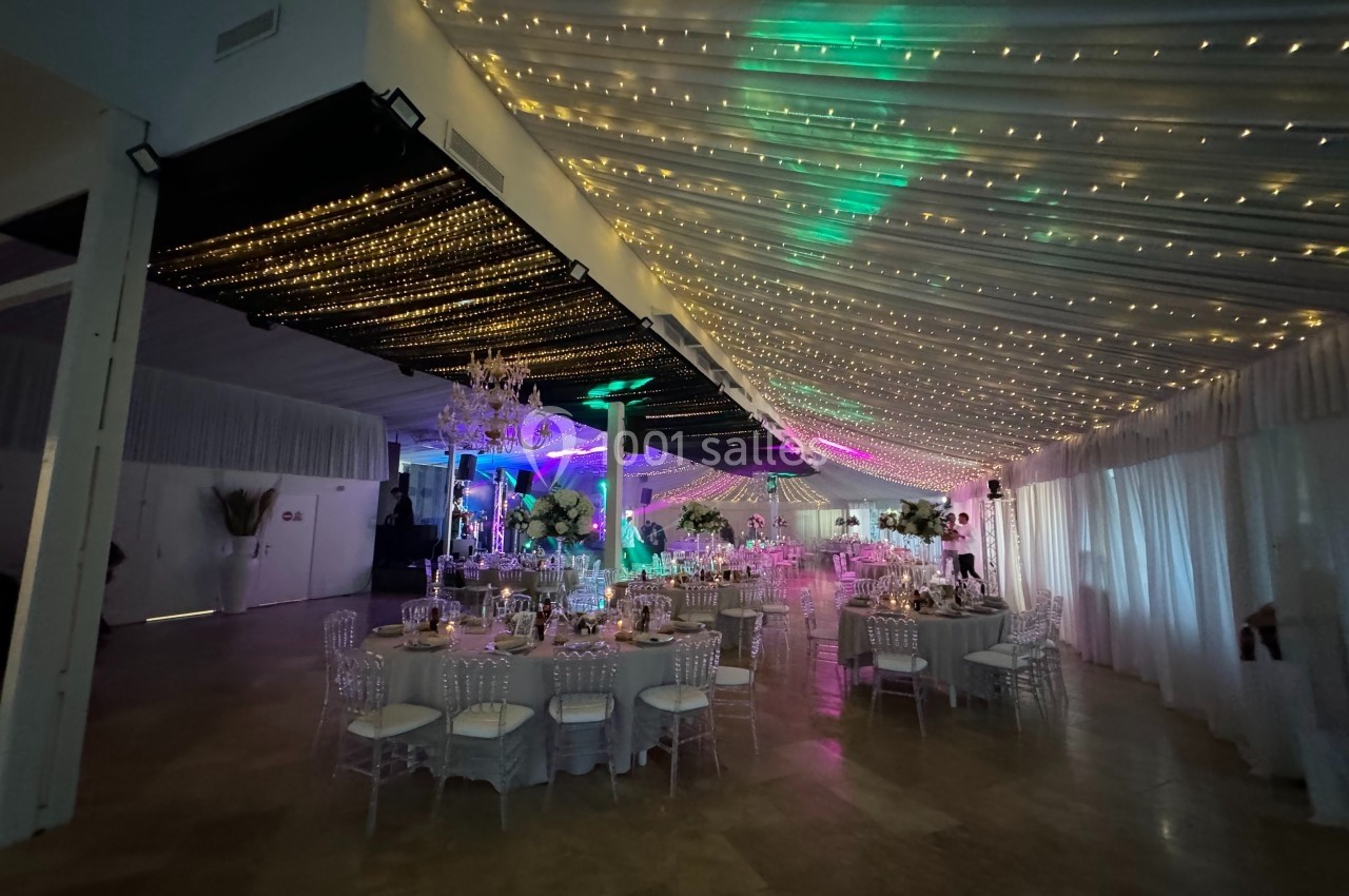 Salle de réception décorée avec des guirlandes lumineuses suspendues, tables rondes dressées et éclairage coloré.