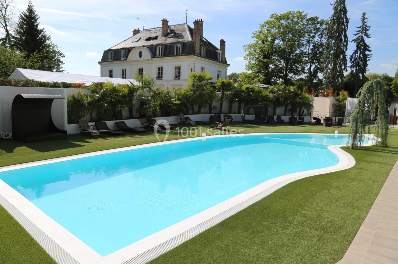 Piscine extérieure entourée de chaises longues, pelouse artificielle et végétation, avec un bâtiment en arrière-plan.