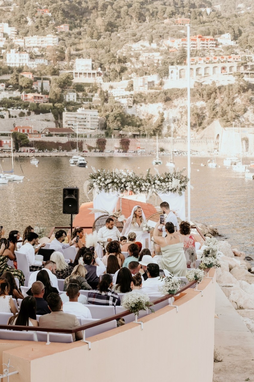 Cérémonie de mariage en plein air au bord de l'eau, avec des invités assis et un paysage urbain en arrière-plan.
