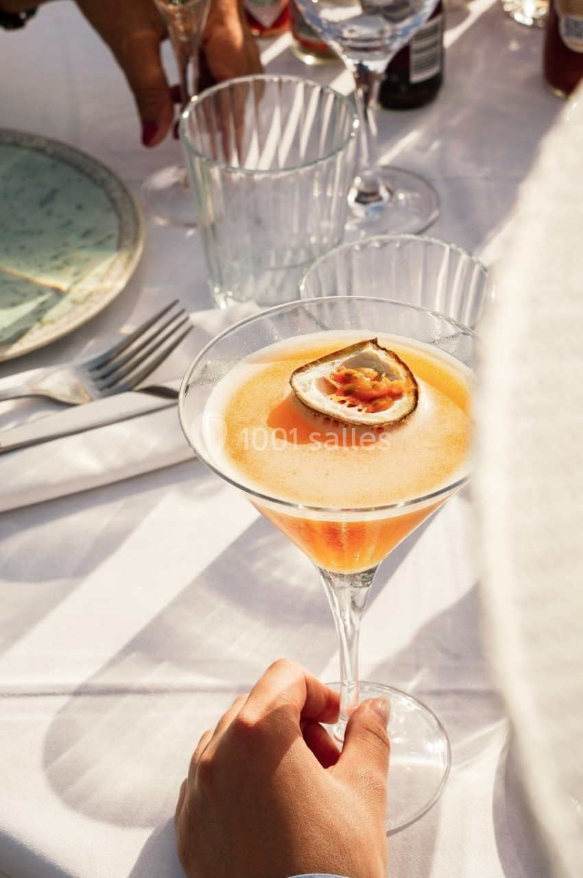 Un cocktail orange garni d'un demi-fruit de la passion, tenu par une main sur une table dressée en extérieur.