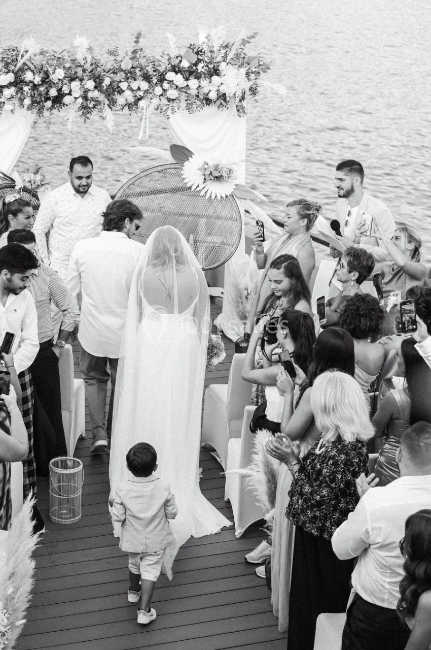 Une mariée en robe blanche avance vers un autel fleuri au bord de l'eau, entourée d'invités prenant des photos.