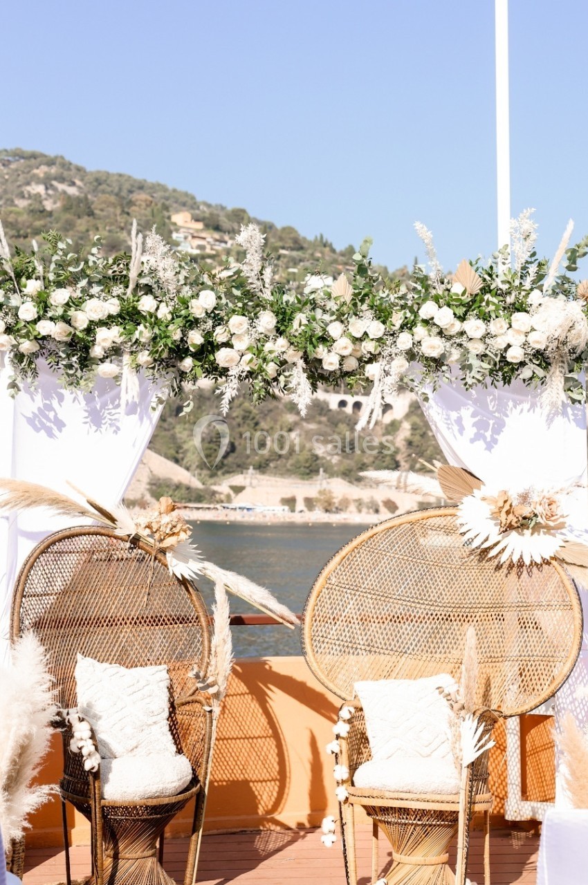 Deux fauteuils en rotin décorés de fleurs séchées sous une arche fleurie, avec vue sur un paysage côtier en arrière-plan.