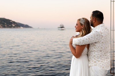 Un couple en tenue élégante regarde la mer au coucher du soleil, avec un bateau visible à l'horizon.