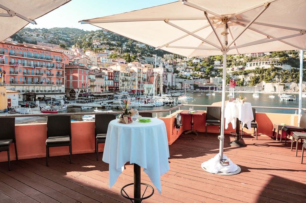 Terrasse ensoleillée avec tables hautes, vue sur un port de plaisance et des bâtiments colorés en bord de mer.