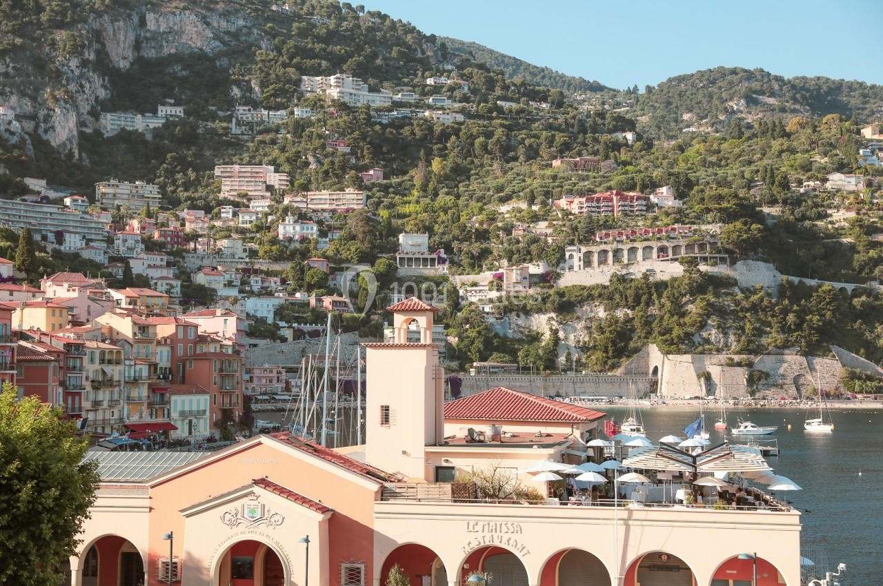 Vue sur un port méditerranéen avec des bâtiments colorés, des collines verdoyantes et des bateaux amarrés.