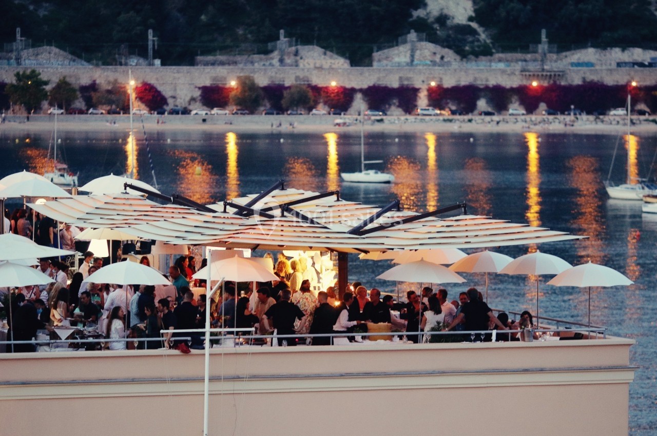 Terrasse animée d'un restaurant en soirée, avec vue sur une baie éclairée et des bateaux au mouillage.