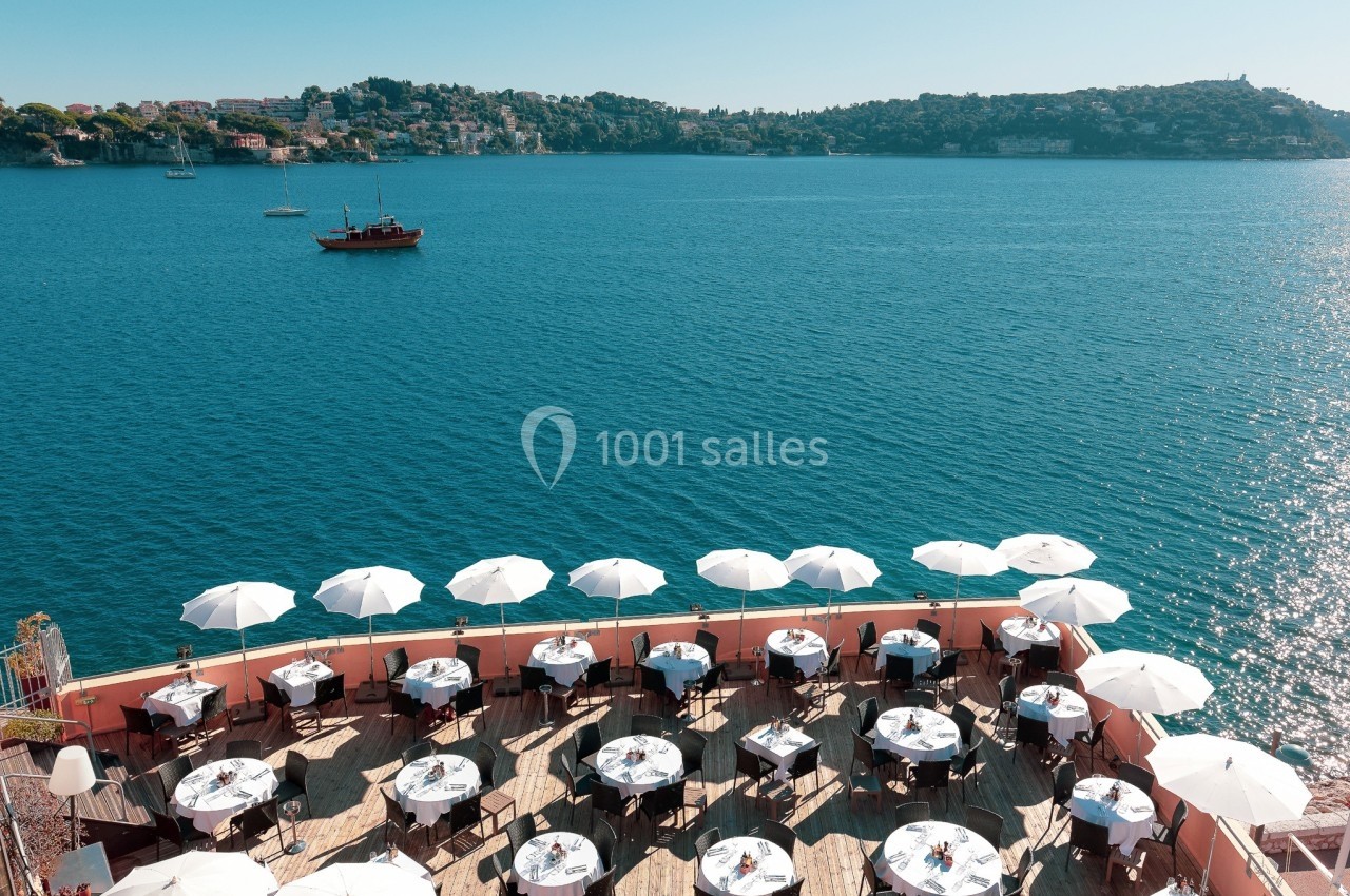 Terrasse d'un restaurant en bord de mer avec des tables dressées et des parasols blancs, vue sur une baie et des collines.