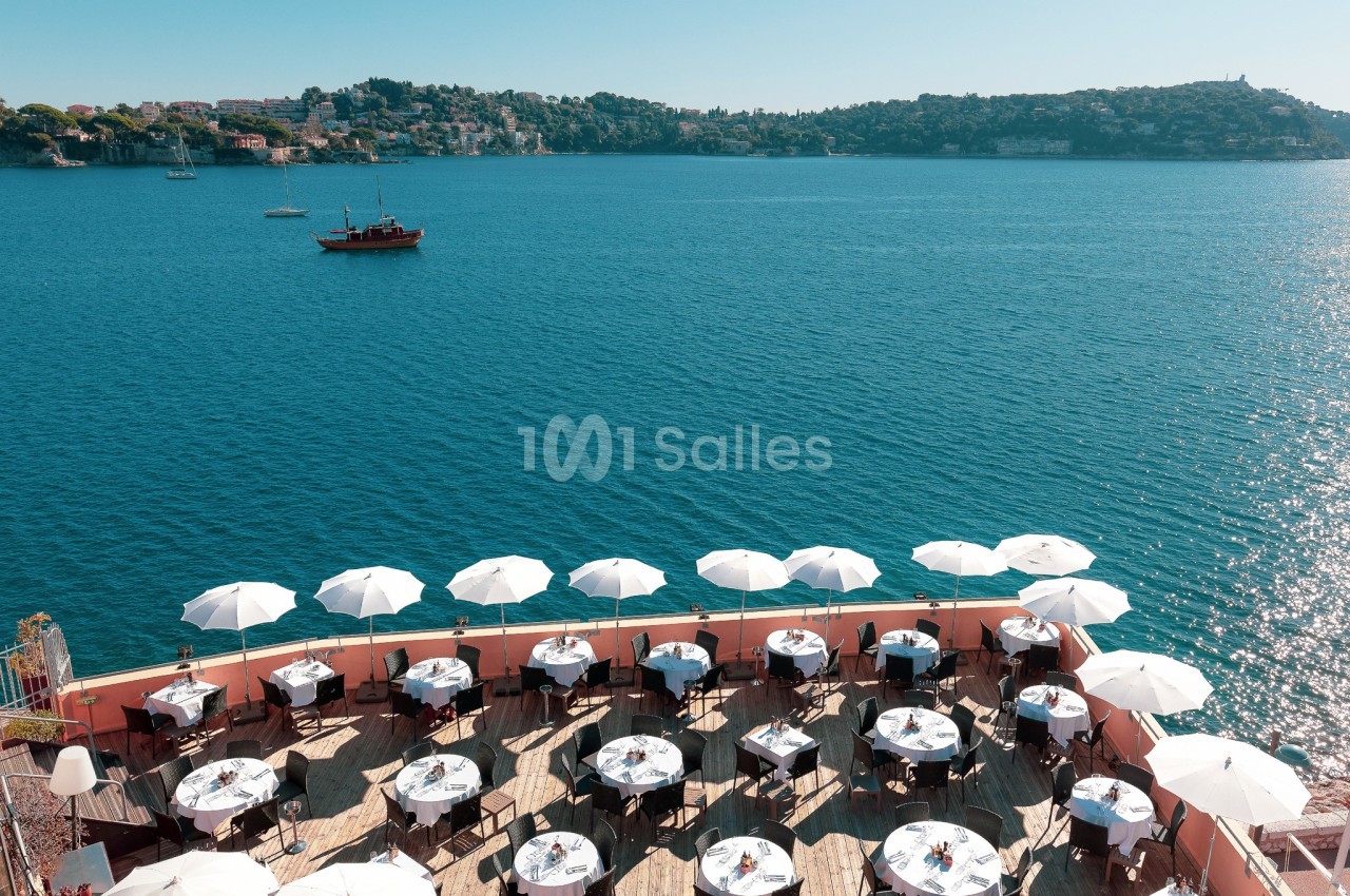 Terrasse d'un restaurant en bord de mer avec des tables dressées et des parasols blancs, vue sur une baie et des collines.