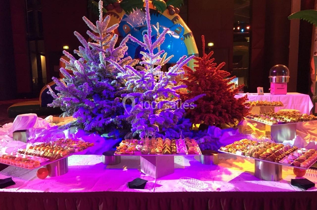 Buffet de desserts présenté devant des sapins décoratifs colorés, éclairés par des lumières vives dans une salle festive.