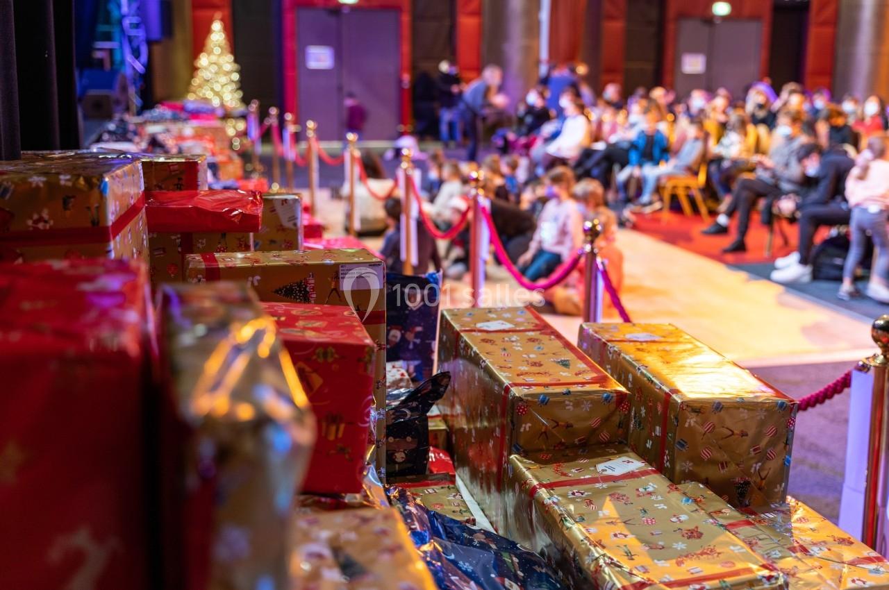 Des cadeaux emballés sont empilés au premier plan, avec un public assis en arrière-plan dans une salle décorée.