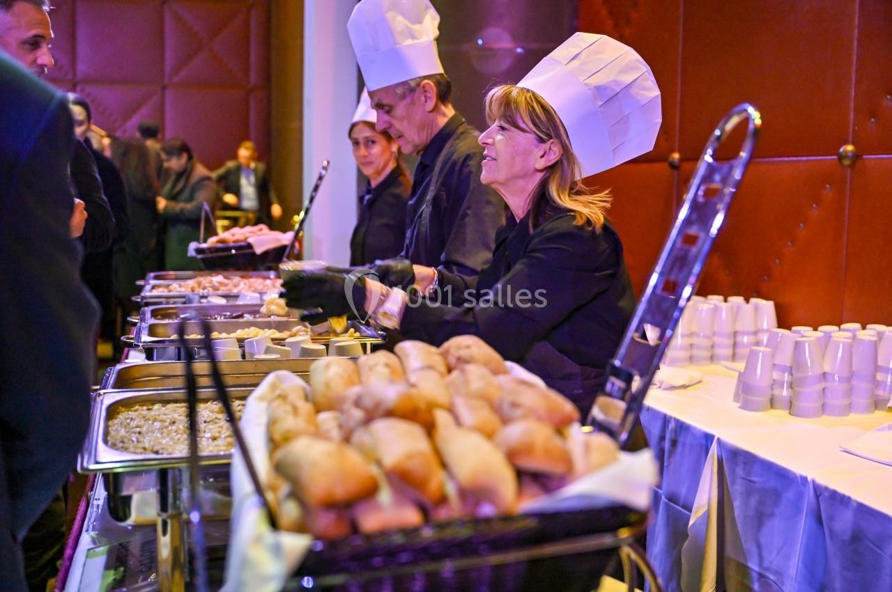 Des chefs en tenue servent des plats variés sur un buffet, avec des pains en premier plan et des convives en arrière-plan.