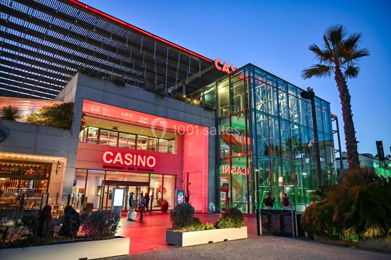 Façade d'un casino moderne avec enseigne lumineuse rouge, entrée vitrée et végétation en premier plan au crépuscule.