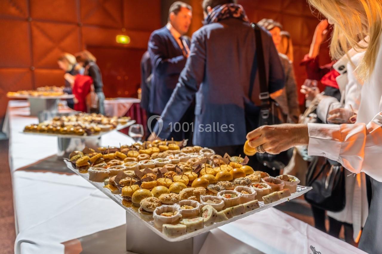 Plateaux de bouchées apéritives variées sur une table lors d'un événement avec des invités en arrière-plan.