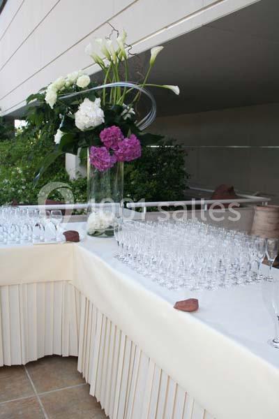 Table dressée avec nappes blanches, verres alignés et grand vase contenant des fleurs blanches et violettes.