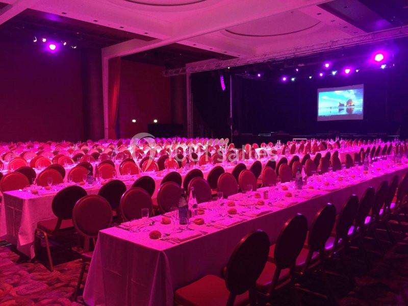 Salle de réception avec tables dressées et éclairage violet, face à une scène avec un écran de projection.
